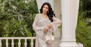 Woman in a white and pink traditional outfit standing outdoors with greenery and architectural elements.